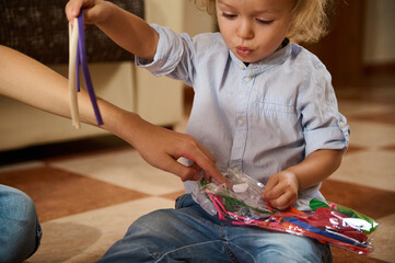 A toddler sits on a checkered floor opening a colorful package of craft supplies, showing curiosity...