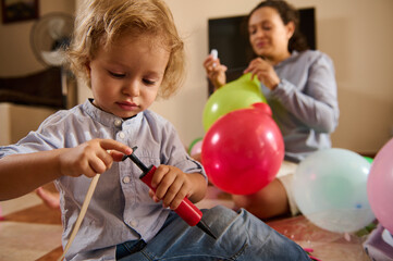 A young child concentrates on inflating a balloon with a manual pump, assisted by a cheerful parent...