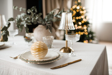 Kitchen table with dinner christmas set over xmas tree with glowing lights in room close up. Winter holiday season.