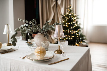 Kitchen table with dinner christmas set over xmas tree with glowing lights in room close up. Winter holiday season.