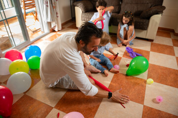 Fototapeta premium A father and his young children engage in balloon crafting in a vibrant room. The scene embodies family bonding, creativity, and fun, surrounded by colorful balloons enhancing the joyful atmosphere.