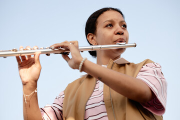Young adult woman playing flute outdoors, holding instrument with both hands, looking slightly...