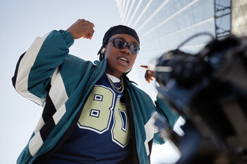 Black young adult man wearing sunglasses gesturing confidently outdoors, standing in urban...
