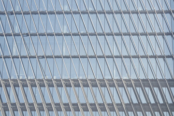 Modern glass building facade featuring repeating vertical and horizontal lines reflecting sky, architectural detail emphasizing geometric patterns and transparency, no people visible