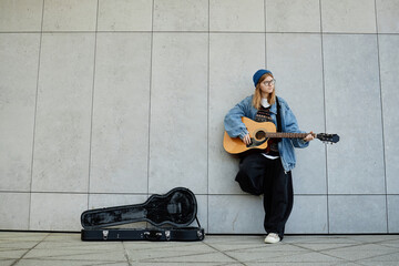 Caucasian teenager girl standing against tiled wall playing acoustic guitar, wearing glasses and...