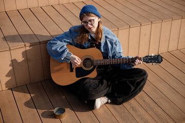 Caucasian teenager girl sitting on wooden steps playing acoustic guitar outdoors, wearing glasses...