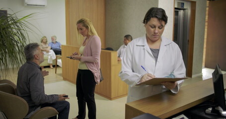 Obraz premium Female doctor reviewing clipboard notes at clinic reception desk while patients wait in background and staff engage with visitors in busy medical lobby