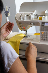 A woman meticulously sews bright yellow fabric on a modern, white sewing machine. Her hands...