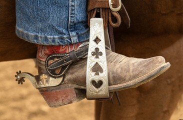 A closeup view of a custom designed silver stirrup with a colorful leather western style boot and a spur.