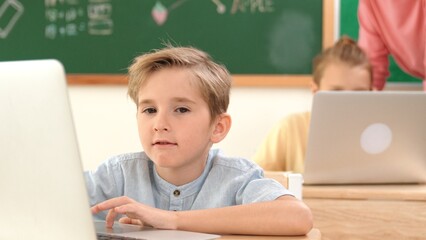 Attractive caucasian boy looking at camera while studying math at school. Diverse cute student doing test while teacher asking asian child question and point at paper in front of blackboard. Pedagogy.