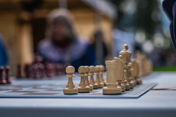 White chess pieces lined up at the start of a game on an outdoor board, with players blurred in the...