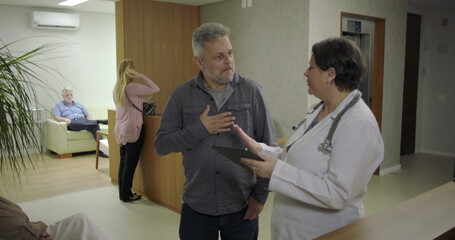 Obraz premium Man expressing relief while speaking with female doctor in clinic hallway, smiling with hand on chest as he receives reassuring news during healthcare discussion