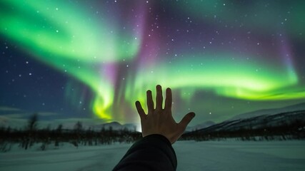 A hand reaches towards the vibrant aurora borealis dancing across the night sky, illuminating the snowy landscape. - Powered by Adobe