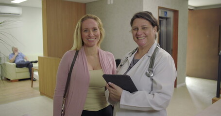 Obraz premium Smiling doctor and patient standing side by side in clinic hallway, holding digital tablet and looking at camera in bright healthcare environment