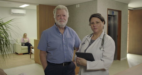 Obraz premium Senior man and female doctor standing together in clinic hallway, facing camera with calm and serious expressions, holding tablet in professional setting