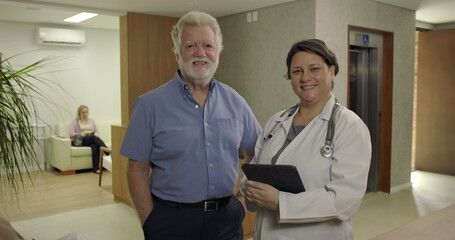 Obraz premium Senior male patient and female doctor smiling together in clinic hallway, standing side by side near reception with digital tablet in hand