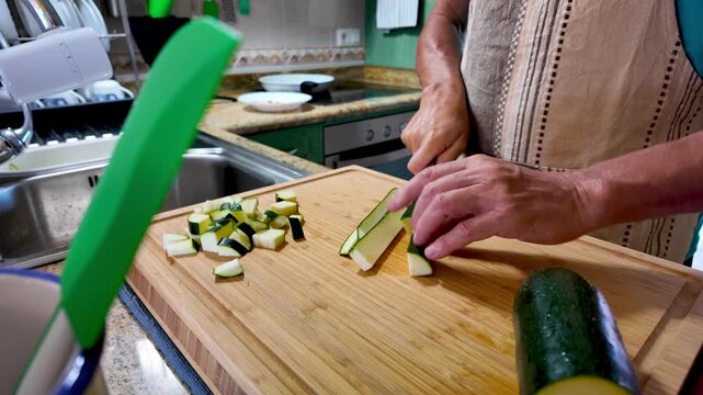 Senior chef wearing an apron slices zucchini on a wooden cutting board in a home kitchen, preparing ingredients for pasta with ham, zucchini and parmesan