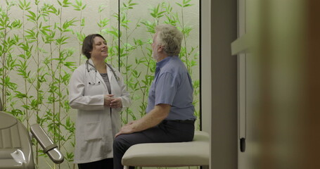 Fototapeta premium Senior male patient entering clinic room and approaching smiling female doctor before sitting on examination chair in modern healthcare facility