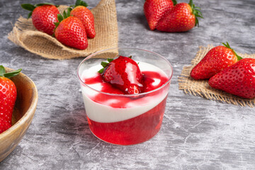 Glass cup with strawberry jelly, cream, and fresh strawberry on top, placed on a dark slate surface with whole strawberries around.