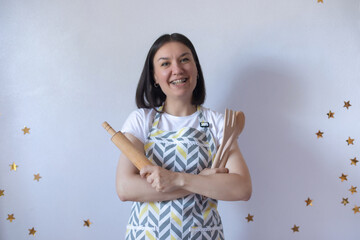 Smiling young woman with braces wearing a patterned apron holds a rolling pin and wooden utensils,...