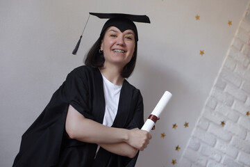Joyful young woman in graduation cap and gown holds diploma, smiling proudly against a decorated...
