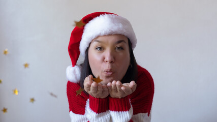 Smiling young woman in a Santa hat and red striped sweater blows golden star confetti from her hands, creating a joyful festive holiday atmosphere against a white background.