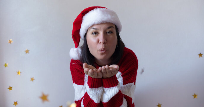Smiling young woman in a Santa hat and red striped sweater blows a kiss toward the camera against a festive background, spreading holiday cheer and warmth. - Powered by Adobe