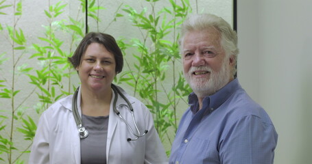 Female doctor and elderly male patient smiling together in clinic setting with green plants in background, showcasing positive medical relationship