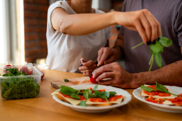 Close-up of couple's hands working together at wooden counter, slicing tomatoes and arranging fresh spinach leaves for morning sandwiches.