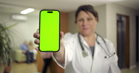Doctor presenting smartphone with green screen in hospital hallway while people wait near reception and elevator in background