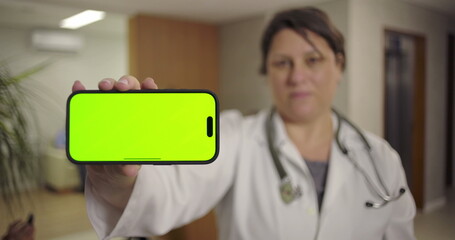 Doctor presenting smartphone with horizontal green screen in hospital lobby with blurred background of waiting area and elevator