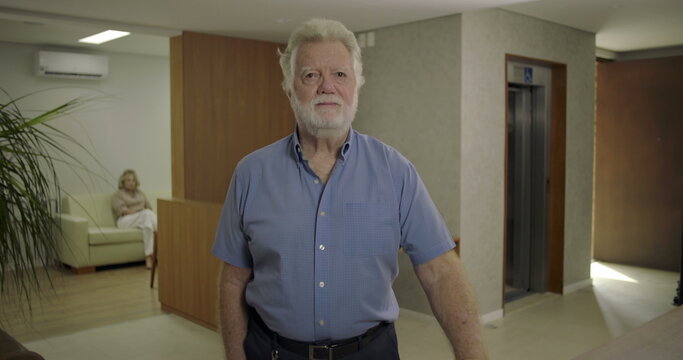 Senior man standing in hospital lobby with woman seated in background as doctor walks into elevator