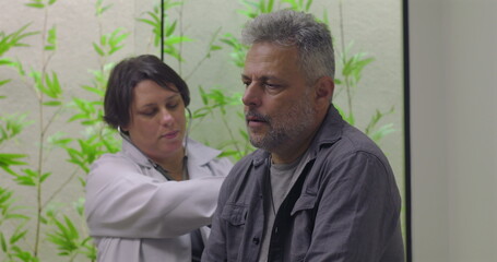 Female doctor conducting chest examination with stethoscope on middle-aged male patient in clinic setting featuring green foliage wall