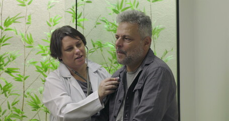 Doctor using stethoscope on chest of middle-aged male patient during medical examination in clinical environment with leafy background