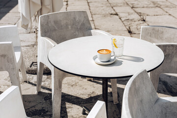Cappuccino and a glass of water with lemon on the outdoor table surrounded by the concrete chairs.