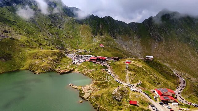 Flying over the green mountain lake with touristic infrastructure nearby. Lots of people and cars are near the waterscape. Breath-taking scenery of Romania.