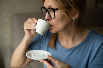 Happy dreamy middle aged woman is staying in living room with cup of black tea or coffee, looking away, Peaceful mature lady enjoying no stress calm positive pastime alone at home.
