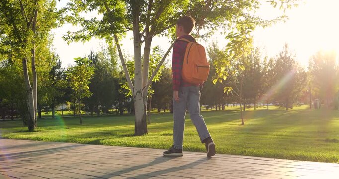 Happy child boy with backpack walks along park path on sunny day. Kid in plaid shirt and jeans enjoys nature, golden sunset light and peaceful walk. Concept of childhood, school and outdoor leisure. 