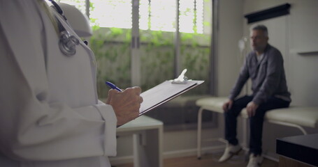 Closeup of female doctor holding clipboard and pen as middle-aged male patient waits in exam room background