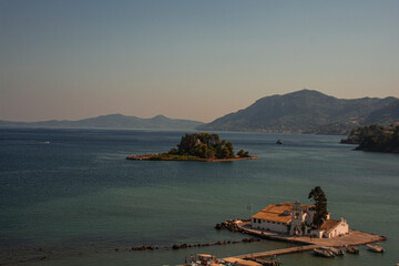 Vlacherna Monastery, located on a small islet near Corfu, Greece, is seen in the foreground with its iconic white buildings and red-tiled roof. The calm blue waters of the Ionian Sea surround it. In t