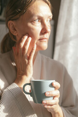 Mature caucasian woman is drinking tea or coffee from cup near window at home. Concept of domestic lifestyle. Cropped photo of modern happy middle aged lady wearing bathrobe at sunny day.