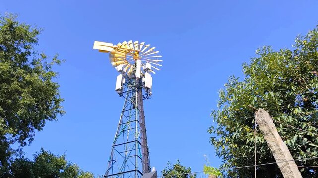 Cata-vento com estrutura de telecomunica&ccedil;&otilde;es em meio a algumas &aacute;rvores e um c&eacute;u azul