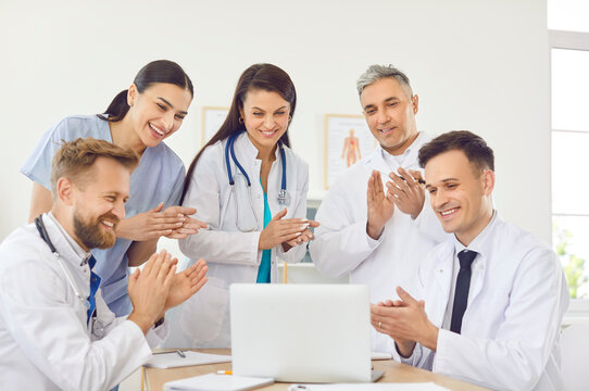 Happy smiling doctors rejoicing at success in treatment applauding standing at desk in clinic during meeting looking at laptop. Successful team of young medical staff in hospital. Health care concept