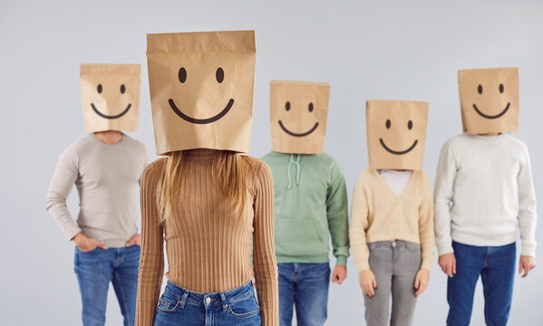 Person with paper bag over head with happy emoticon mouth and eyes facial expression drawn on it. Casual young woman in smiling package bag standing in gray studio with group of people in background