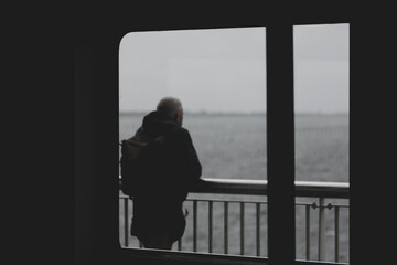 Selective focus of boat window frame with rain and cloudy in dark tone, Silhouette of a man standing on balcony of the ferry looking outside view, Blurred passenger in public transportation background