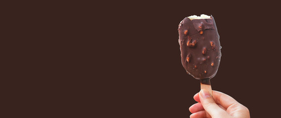 Chocolate covered ice cream bar on Dark brown background, during the holidays. Hand holds delicious chocolate and nuts almond ice cream bar on a stick.