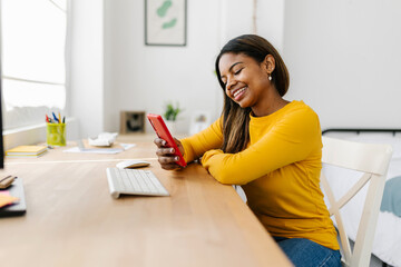 Happy young african american woman using smart phone while working remotely from home office