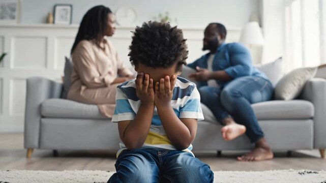 sad little african american boy covered his face with his hands, parents arguing nearby