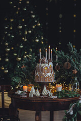 Christmas and New Year beautiful elegant family dinner table setting decorated with candles, fir tree branches, handmade craft toys. Gorgeous big homemade cake with gingerbread houses as centrepiece.