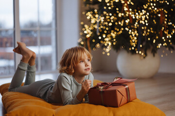 Happy Caucasian girl, 9 years old, blond curly hair, in pajamas, lying on soft cushion holding gift...